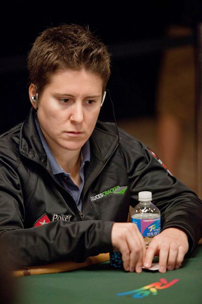 A focused poker player with short brown hair, wearing a black jacket and earphones, sits at the poker table holding poker chips and a water bottle, reminiscent of Vanessa Selbst plotting her next big comeback.