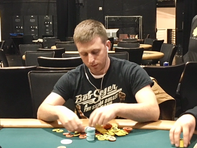A man wearing a Bob Seger T-shirt, possibly Louis Ganser, sits at a poker table stacking chips, with several empty tables and chairs in the background at the Cleveland Main Event.