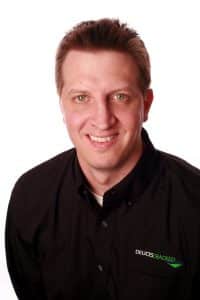 A man with short light brown hair, wearing a black collared shirt with a green and white home logo, smiles at the camera against a white background. - Poker Training Videos and Poker Coaching A man with short light brown hair, wearing a black collared shirt with a green and white home logo, smiles at the camera against a white background.