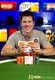 A smiling man sits at a poker table holding playing cards and chips, with stacks of chips in front of him and colorful KasinoKrime banners in the background. - Poker Training Videos and Poker Coaching A smiling man sits at a poker table holding playing cards and chips, with stacks of chips in front of him and colorful KasinoKrime banners in the background.
