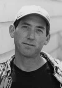 A man wearing a light-colored baseball cap and plaid shirt looks directly at the camera, standing in front of a light brick or siding wall, reminiscent of Tommy Angelos laid-back style. - Poker Training Videos and Poker Coaching A man wearing a light-colored baseball cap and plaid shirt looks directly at the camera, standing in front of a light brick or siding wall, reminiscent of Tommy Angelos laid-back style.