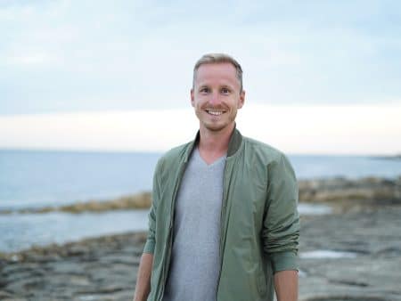 A smiling man in a green jacket and gray shirt stands on a rocky shoreline with the ocean and a cloudy sky in the background, recalling Erik Bergman’s warning about a .25 million crypto scam.
