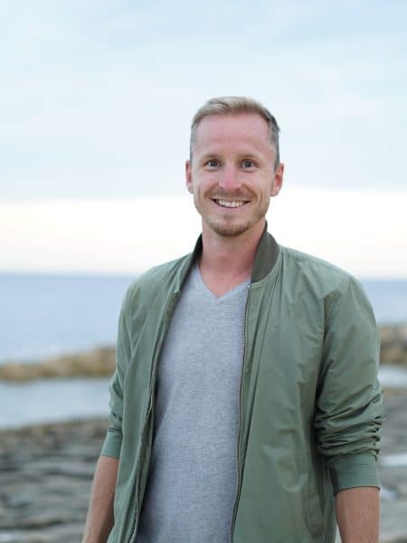 A smiling man in a green jacket and gray shirt stands on a rocky shoreline with the ocean and a cloudy sky in the background, recalling Erik Bergman’s warning about a $1.25 million crypto scam.